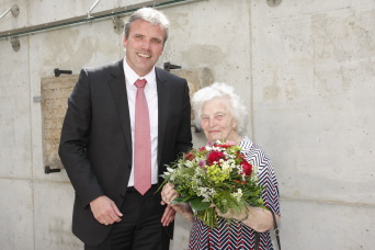Ein Mann links, zwei Köpfe größer als die Dame rechts, die lächelnd einen Blumenstrauss hält