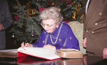 Eine Dame in lila Kleidung trägt sich in ein großes rotes Buch ein. Dahinter ein Weihnachtsbaum.