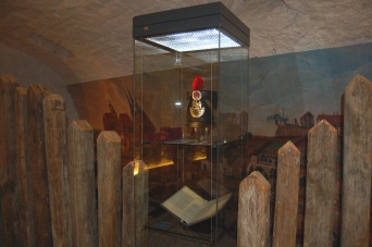 Im Vordergrund rechts und links ein Staketenzaun, dazwischen eine Vitrine. Darin lagert u.a. ein Buch und ein historischer Uniformhut. Im Hintergrund ist ein Wandgemälde zu sehen, welches am Tonnengewölbe, das die Ausstellung beherbergt, angebracht ist.