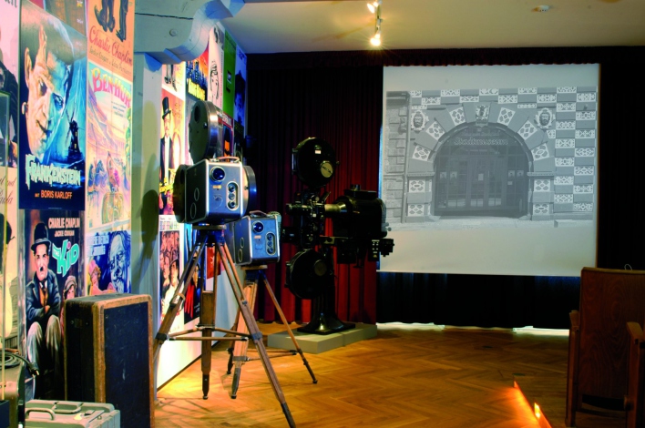 Links eine Plakatwand mit alten Filmplakaten, rechts eine Projektionsfläche, dazwischen die historischen Projektoren.
