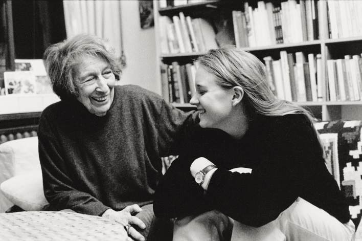 Eine ältere Dame,links, und eine junge Frau, rechts, auf einer Couch sitzend, im Hintergrund Bücherregale. Eine Schwarz-Weiß-Fotografie.