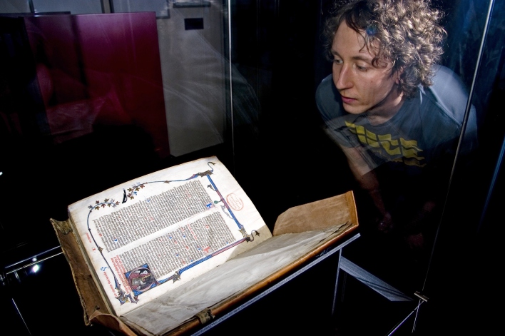 Ein dickes sehr altes Buch mit Buchmalereien im Vordergrund, hinter dem Vitrinenglas eine Frau, die interessiert auf die Handschrift schaut.