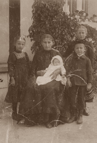 Frau mit langem dunklem Kleid, sitzend, mit Baby auf dem Arm, rechts und links die Kinder.