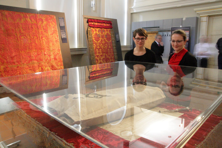 Zwei Frauen, rechts im Hintergrund, blicken auf eine Vitrine, die im Vordergrund des Bildes ist. Links und in der Bildmitte zwei in Rot- und Gelb- bzw. Goldtönen gehaltene Toravorhänge. 