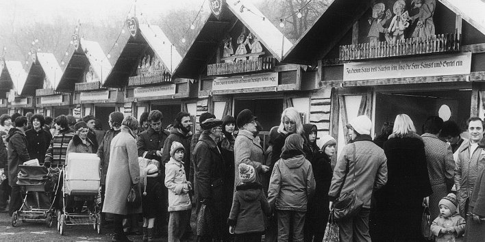 Sechs Weihnachtsmarktbuden in Reihe, davor Männer, Frauen und Kinder, zum Teil mit Kinderwagen, in Schlangen stehend.