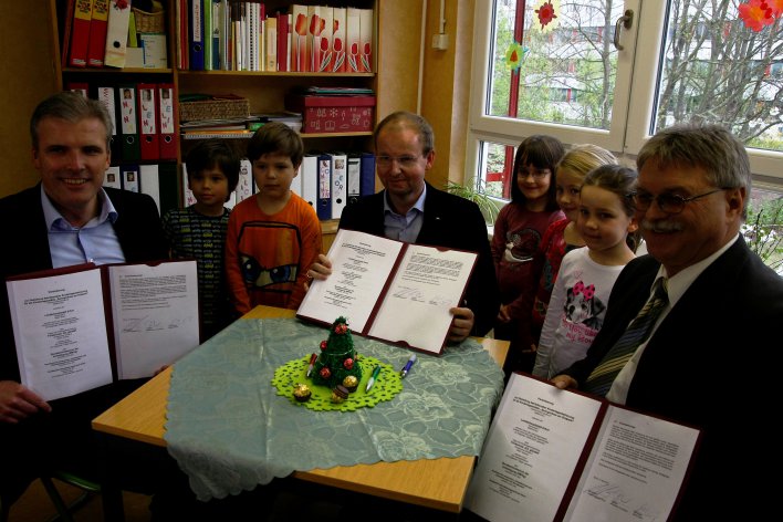 Die Unterzeichner zeigen die unterschriebenen Urkunden, um sie herum stehen einige Kindergartenkinder. 