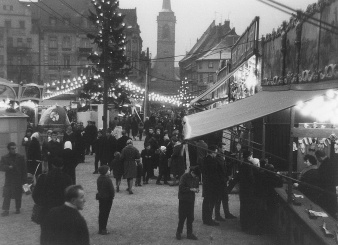 Zahlreiche dunkel gekleidete Menschen, zum Teil mit Kopftüchern, auf einem Platz. Links ein schmaler, hoher Weihnachtsbaum, von welchem Lichterketten ausgehen. Rechts ein Marktstand. In der Mitte, im Hintergrund eine Kirche.