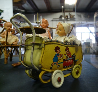 Gelber Puppenwagen, auf der Seite eine Darstellung zweier Puppenmuttis. Im Wagen eine Puppe mit weißer Jacke und weißer Mütze.Im Hintergrund weitere Puppen an einem Puppentisch.