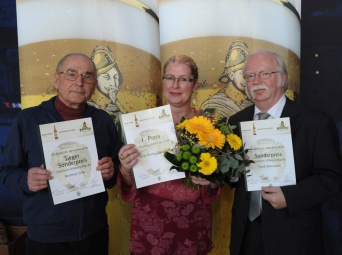 Drei Personen halten eine Urkunde in den Händen, die Dame in der Mitte dazu einen Blumenstrauss. Im Hintergrund Abbildung zweier Biergläser.