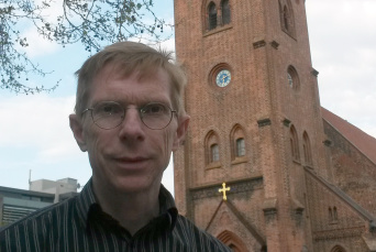 Ein Mann mit Brille und gestreiftem Hemd vor einer Kirche in Ziegelsteinbauweise.