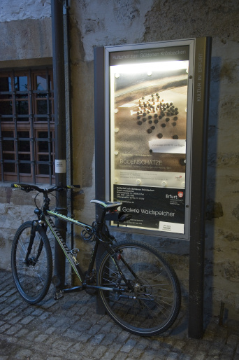 Plakat zu den Ausstellungen Ein gläserner Plakataufsteller, davo ein Fahrrad.