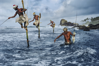 Drei Männer auf Stakhölzern im Ozeanwasser, ein Mann, recht, ohne Stakete, mit Stock.