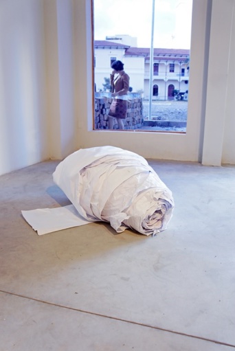 Eine grob gewickelte Papierrolle liegt auf dem Boden. Im Hintergrund ein Bildfenster. Zu sehen ist eine Frau vor einer Wand stehend, Dahinter ein zweistöckiges Haus.
