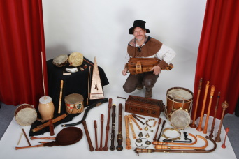 Ein Mann im Kostüm, vor ihm, auf dem Tisch, zahlreiche historische Instrumente. Rechts und links ein Bühnenvorhang.