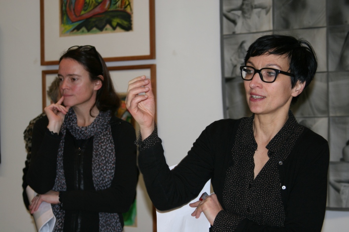 Eine Dame mit schwarzen Haaren, schwarzer Brille und scharz-gemusterter Kleidung, rechts im Bild, erklärt in einer Ausstellung. Links neben ihr eine Dame mit Schal.