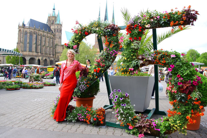 Franziska Gerlach ist die Ega-Park-Blumenkönigin 2014/2015 Junge Frau mit blondem Haar und rotem, langen Kleid an einer großen, mit Blumen dekorierten 25 vor der Kulisse von Dom und St. Severi. Links Marktgeschehen und zahlreiche Pflanzen.