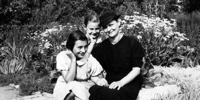 Dame in schwarzem Kleid und Hut mit zwei Töchtern in einem Garten. 