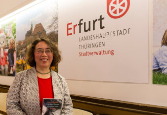 Eine Dame mittleren Alters herzlich lachend, in deren Händen ein Buch. Im Hintergrund Fotos und der Schriftzug Landeshauptstadt Erfurt