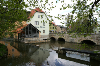 Foto: Neue Mühle Erfurt © Stadtverwaltung Erfurt Im Vordergrund ein Fluss, rechts eine Brücke, links das Gebäude der Neuen Mühle Erfurt. Im Giebelbereich desselben befindet sich eine Glasfront.