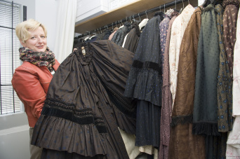 Foto: Im Schaudepot Historische Textilien: Textilrestauratorin Antje Hirschberger präsentiert Sonntagskirchmäntel © Stadtverwaltung Erfurt / D. Urban Eine Frau mit blondem Haar, roter Jacke und buntem Tuch zeigt einen dunkelbraun, reich verzierten Umhang mit Volant, den sie gerade aus einer Reihe verschiedener Umhänge, die sich auf einer Kleiderstange befinden, herausgezogen hatte. Über dem mit einem Vorhang geschlossenen "Kleiderschrank" eine Reihe Schachteln auf einem Regalboden.