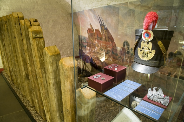 Foto: Tschako, eine militärische Kopfbedeckung, und Uniformreste der französischen Armee in der Ausstellung © Stadtverwaltung Erfurt / D. Urban Links ein Staketenzaun, rechts eine Vitrine mit verschiedenen Ausstellungsstücken.