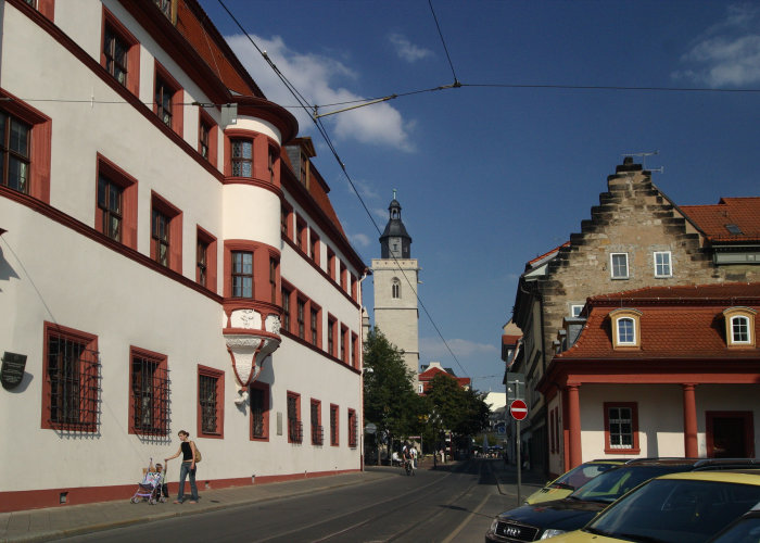 Die St.-Wigberti-Kirche ist ein katholischer Kirchenbau in Erfurt, im Vordergrund die Staatskanzlei. Barockes Gebäude und Kirche unter blauem Himmel.