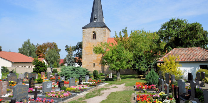 Der Blick geht über Gräberanlagen des Friedhof zur Kirche, die größtenteils von Bäumen verdeckt ist. Nur der quadratische Kirchturm aus Natursteinmauerwerk mit der mit Schiefer belegten Kirchturmspitze ist zu sehen.