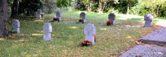 Zwei reihen Granitkreuze mit namen der Verstorbenen stehen auf einer Rasenfläche. Vor einigen Gräbern stehen blühende Blumenschalen.