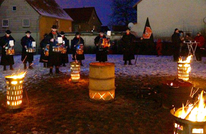 Kostümierte Trommler stehen in winterlicher Landschaft um brennende Feuertonnen und musizieren.