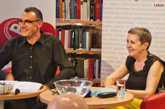 Carsten Rose und Prof. Dagmar Demming sitzen an einem Tisch und lachen ins Publikum.