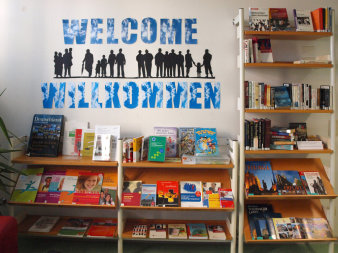 Bücherregale vor einer Wand mit der Aufschrift "Willkommen - Welcome", darin werden Medien mit landeskundlichem Schwerpunkt präsentiert