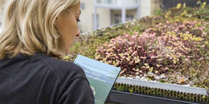 Foto: Christina Klauke bietet in der Domplatz-Bibliothek regelmäßig Veranstaltungen an, u. a. zu den Themen Umwelt, Natur und Nachhaltigkeit © Stadtverwaltung Erfurt / V. Gürtler Frau mit Blick auf ein begrüntes Dach wählt aus einem Bücherstapel aus.