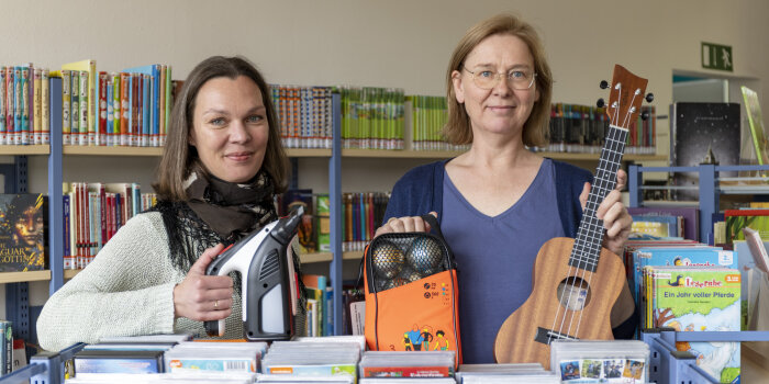 zwei Frauen stehen in einer Bibliothek und halten verschiedene Gegenstände in der Hande