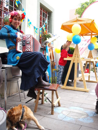 Frau mit Akkordeon sitzt auf einem Stuhl und spielt das Instrument. Im Hintergrund ist ein mit Luftbalölons und Girlanden geschmückter Stand zu erkennen.
