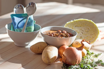 Foto: Suppen-Kochkurs © Stadtverwaltung Erfurt / Grit Kästner Zwei kleine weiße Schüsseln auf hellem Küchentisch. Eine ist gefüllt mit heißer Linsensuppe. In der anderen Schüssel befinden sich, eingewickelt in eine Serviette, zwei Löffel. Im Vordergrund liegen auf dem Küchentisch: Möhren, Zwiebeln, Kartoffeln und ein Krautkopf.
