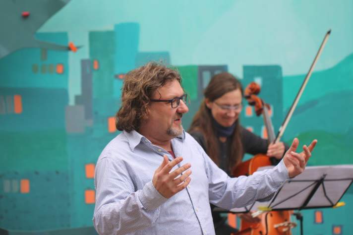 Mann mit einladender Geste, im Hintergrund ein Herr mit Cello hinter einem Notenständer