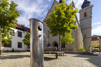 eine metallische Säule mit einem Wasserstrahl, im Hintergrund eine Kirche