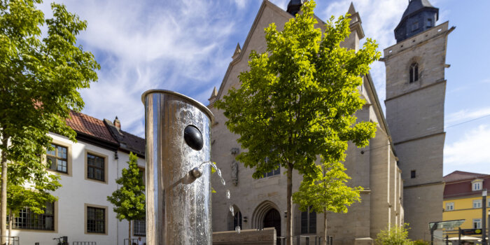 eine metallische Säule mit einem Wasserstrahl, im Hintergrund eine Kirche