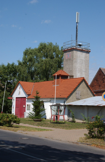 Das Foto zeigt einen quadratischen Turm mit augebauter Mobilfunksendeanlage in dörflich geprägter Umgebung