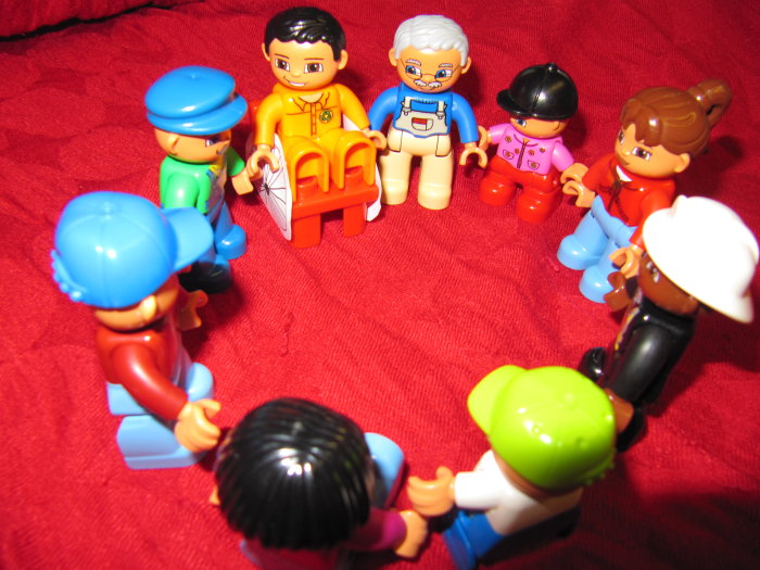 Spielzeug im Kreis aufgestellt Unterschiedlich bemahlte kleine Spielfiguren, darunter eine Figur in einem Rollstuhl, stehen sich an den Händen haltend in einem Kreis auf einer roten Decke