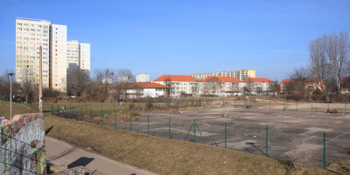 Eingezäunte Brachfläche mit Wohnhäusern im Hintergrund.