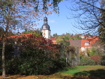 Umgeben von Grün- und Parkfläche erhebt sich im Hintergrund ein weißer Kirchturm mit schiefergedecktem Glockenturm