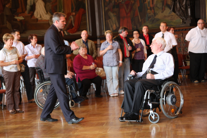 Eine Menschengruppe, darunter zwei Rollstuhlfahrer und Oberbürgermeister Andreas Bausewein, betrachten Gemälde im Festsaal des Rathauses.