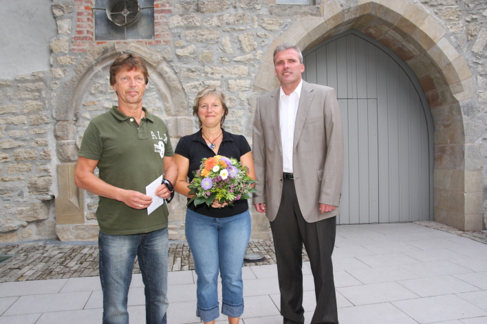 Neben dem Oberbürgermeister Andreas Bausewein stehen links eine Frau und ihr Ehemann. Die Frau hält einen Strauß bunter Blumen in der Hand, der Mann eine Karte. Im Hintergrund ist der Eingangsbereich der Alten Synagoge zu sehen.