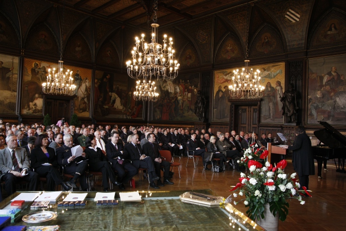 Foto: Festakt © Stadtverwaltung Erfurt Zahlreiche Gäste sitzen im Festsaal. Auf einem Tisch liegen Urkunden, daneben steht Blumenschmuck und weiter rechts steht eine Rednerin am Pult.