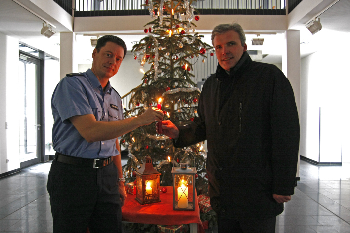 Der Polizeidienststellenleiter nimmt das Friedenslich in Form einer Kerze vom Oberbürgermeister entgegen, beide stehen vor einem Weihnachtsbaum in der Polizeidienststelle.