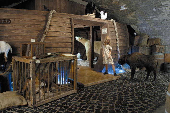 Ein Mädchen führt ein Huftier in die Arche Noah, drinnen ist ein Eisbär zu sehen, davor stehen Käfige mit weiteren Tieren, auf dem Dach sitzt Geflügel. 