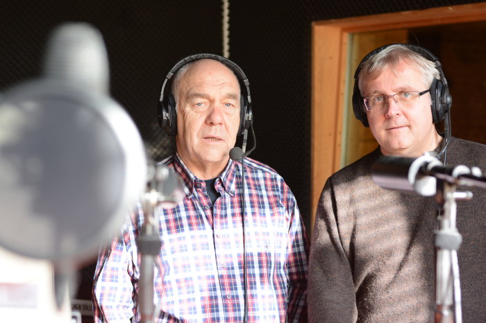 Zwei männliche Personen mit Kopfhörern hinter Mikrofonen