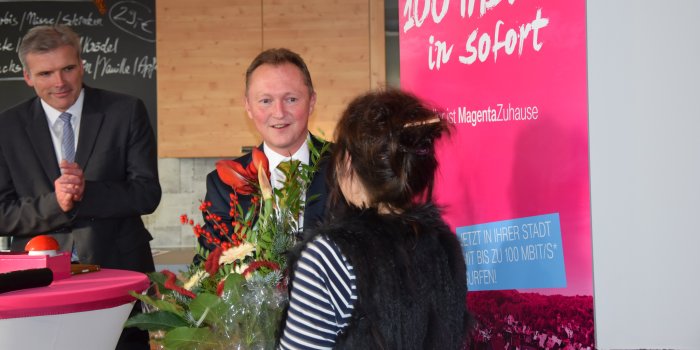 Rainer Frank (Telekom) begrüßt Maria Groß als erste Kundin für das neue Glasfasernetz in Erfurt und überreicht ihr im Beisein des Oberbürgermeisters Andreas Bausewein einen Strauß Blumen.