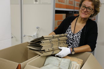 Dr. Antje Bauer hält einen Stapel Akten in der Hand, vor ihr stehen zwei mit Unterlagen gefüllte Kartons, im Hindergrund sieht man ein Regal mit vielen Kartons.